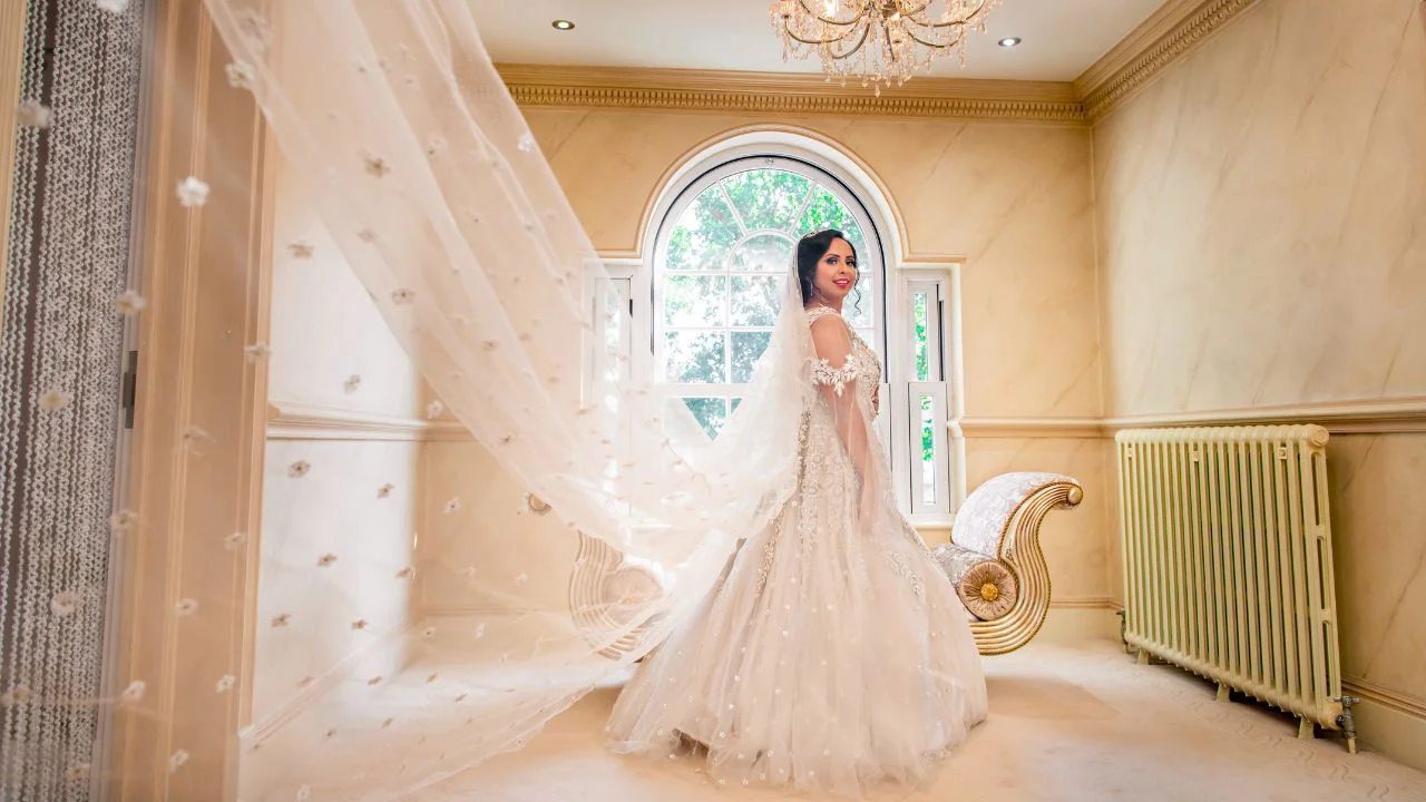 A bride standing in a room with large window in a white dress with a long veil type dupatta smiling.