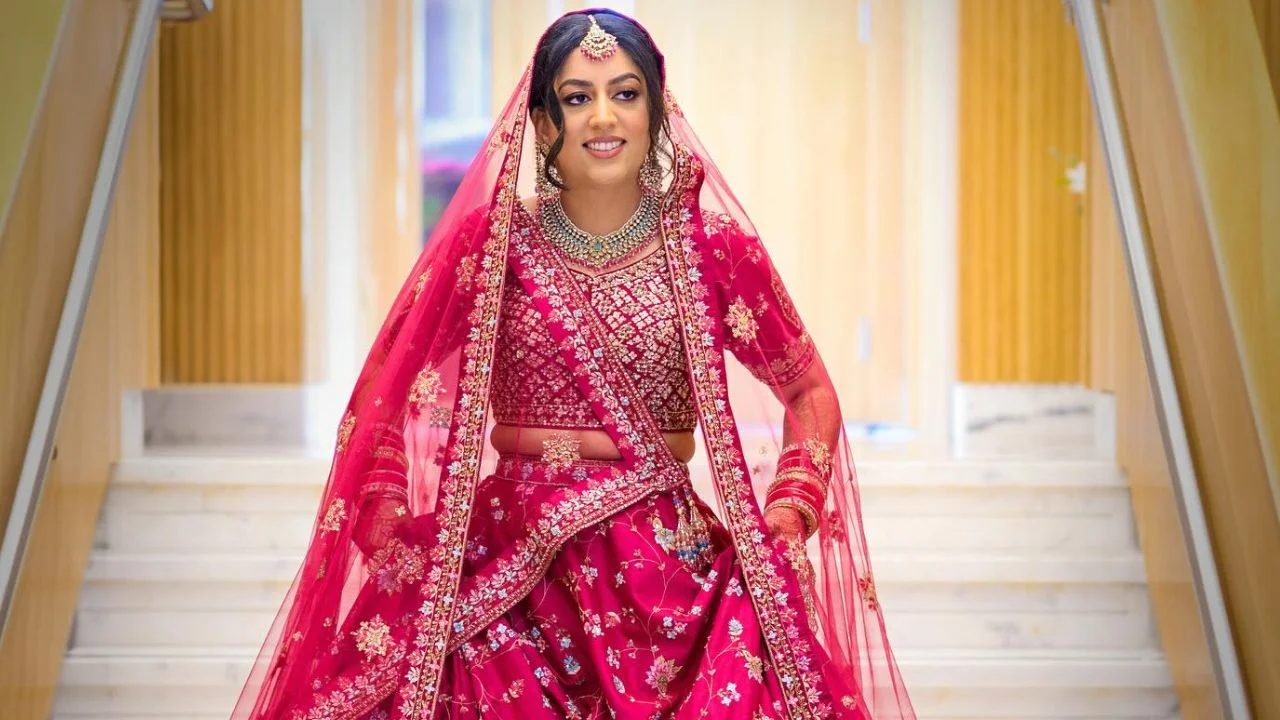 A bride in a pink lengha and dupatta, with a large necklace and earrings coming down through stairs.