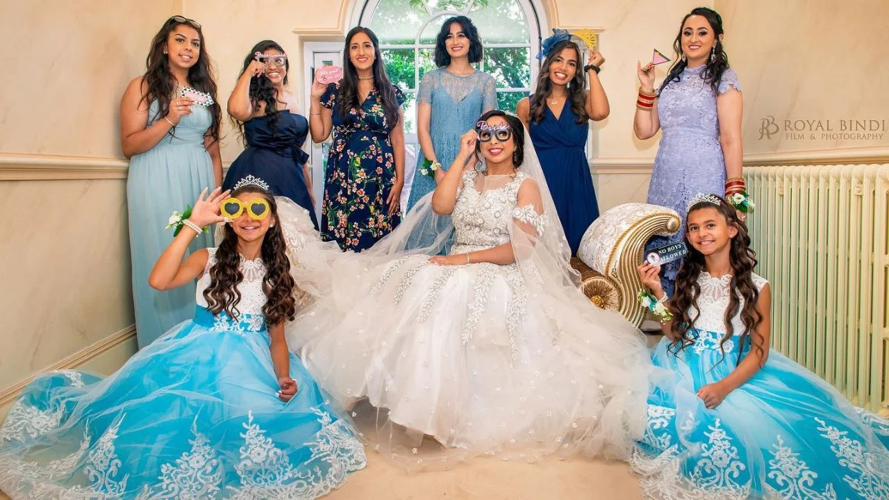 Bride dressed in a white gown with her bridesmaids and flower girls wearing colourful dresses, all posing playfully with fun glasses and props during the pre-wedding celebration.