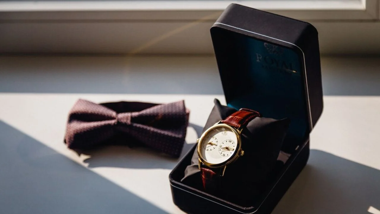 A gold watch in a black box sits next to a purple bowtie on a windowsill. Sunlight casts shadows on the objects.