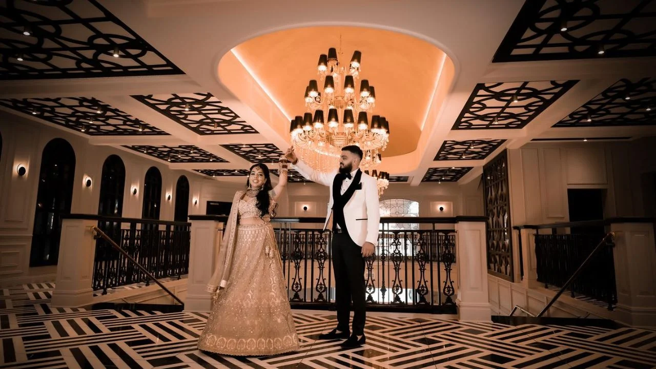 Bride and groom dancing beneath a luxurious chandelier in a grand wedding venue, with intricate ceiling designs and patterned flooring.