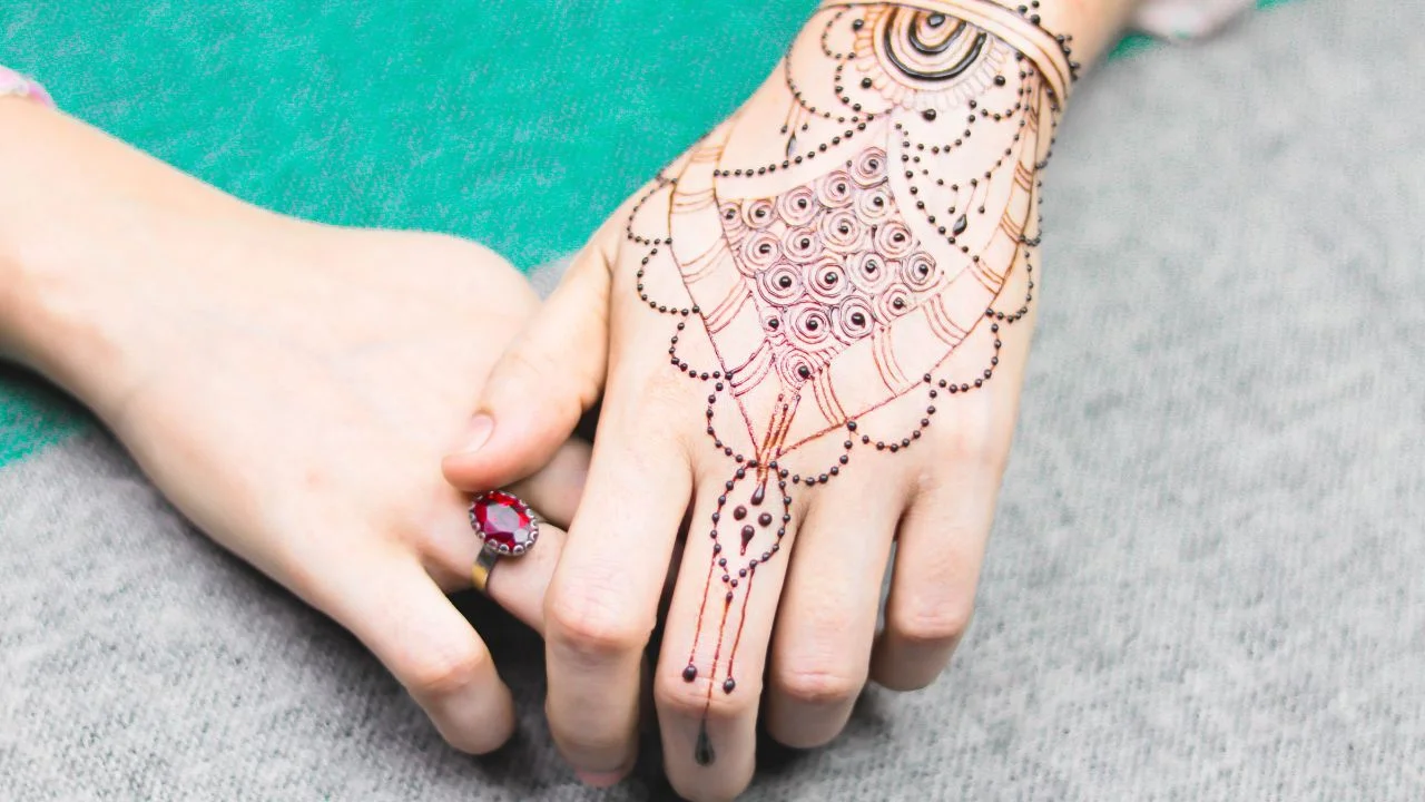 A close-up of a woman's hands with intricate henna designs covers her palm, fingers, and part of her wrist. She is wearing a ring with a red gemstone on her ring finger.