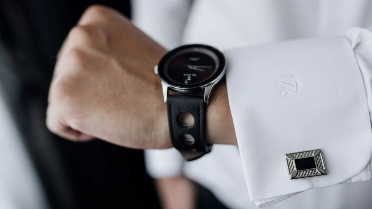 A close-up photo of a man's hand wearing a silver wristwatch with a black leather strap. He is checking the time. He is also wearing a white dress shirt with cufflinks.