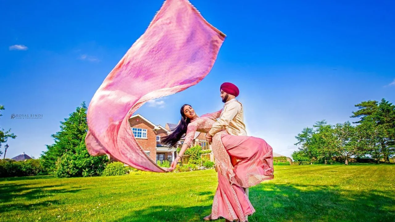 Punjabi couple in traditional wedding attire outdoors, with the bride's pink dupatta flowing dramatically in the air, captured by Royal Bindi Photography