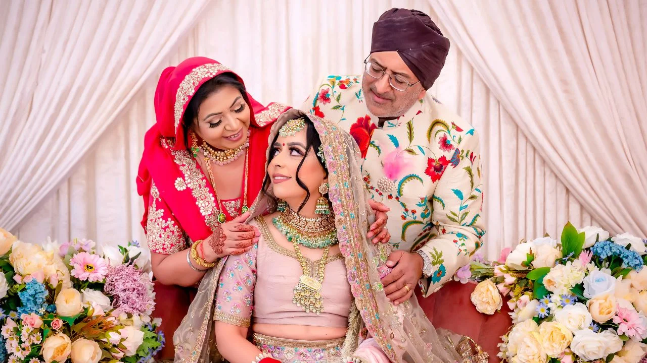 A bride in a pink and gold lengha is being blessed by her parents. She is surrounded by flowers and has a big smile on her face.