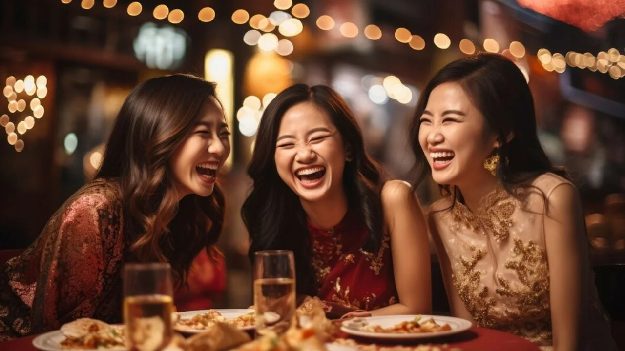 Three women laugh together at a table with plates of food and glasses of wine. The woman on the left has long brown hair and is wearing a red dress, the woman in the middle has long black hair and is wearing a red dress, and the woman on the right has long black hair and is wearing a gold dress.