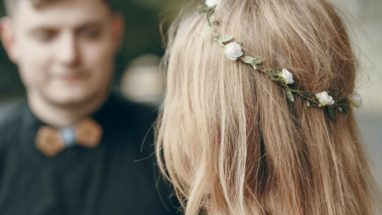 Back view of a woman wearing a delicate floral hair vine, with a man in the background wearing a bow tie.