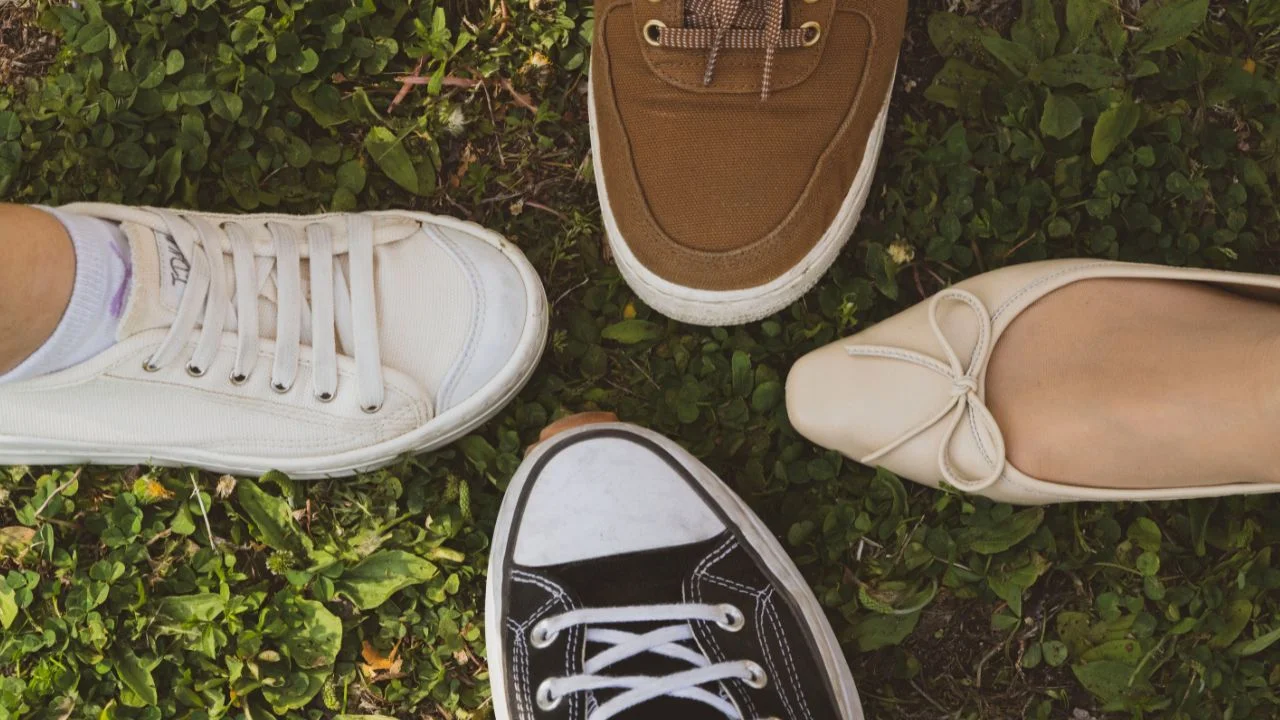 A top-down view of four feet in different shoes: white sneakers, brown lace-up shoes, black Converse sneakers, and beige ballet flats, arranged on a grassy surface.