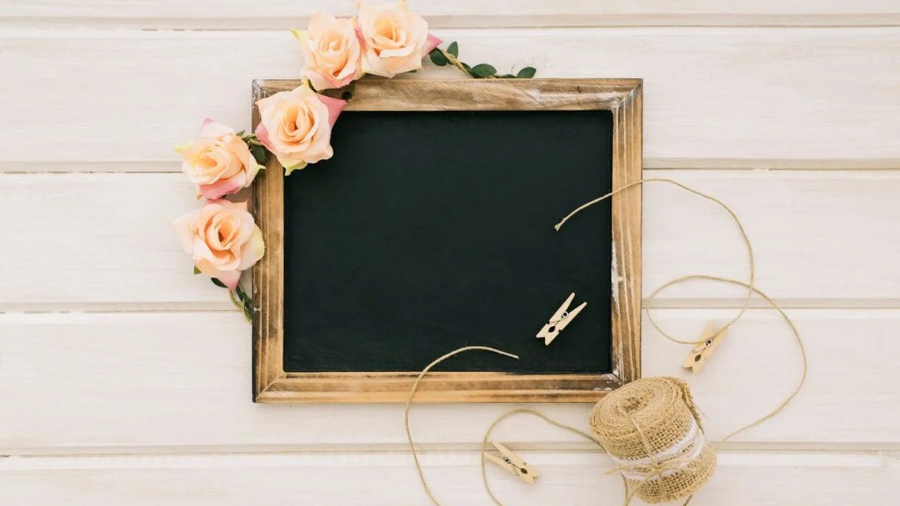 A blank chalkboard surrounded by pink roses, jute twine, and wooden clothespins on a white wooden background.