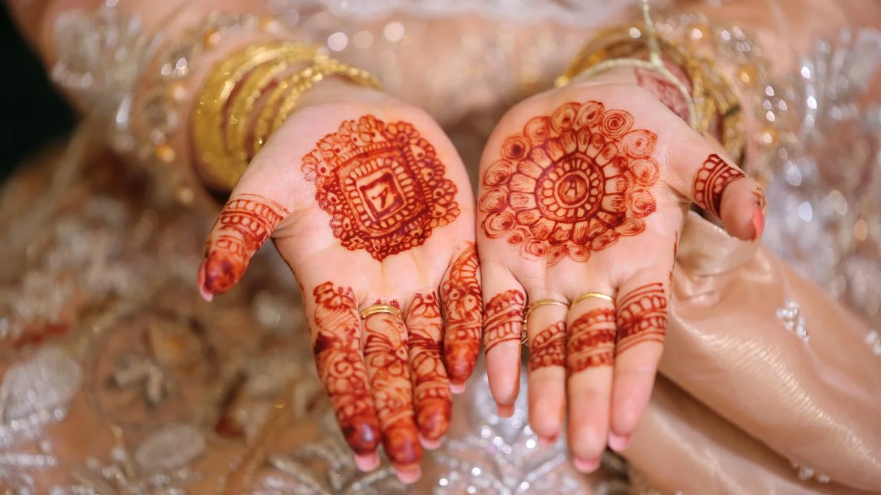 Woman's hands adorned with detailed mehndi designs. The henna is reddish-brown and features circular patterns on the palms. The woman is wearing gold bangles on her wrists and rings on her fingers.