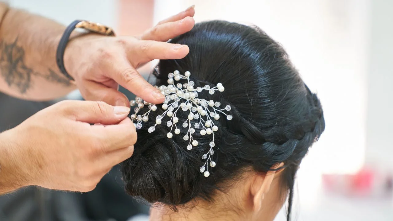 A hairstylist placing a delicate, pearl-embellished hair comb into a dark-haired woman's braided updo, adding an elegant touch to her bridal hairstyle.