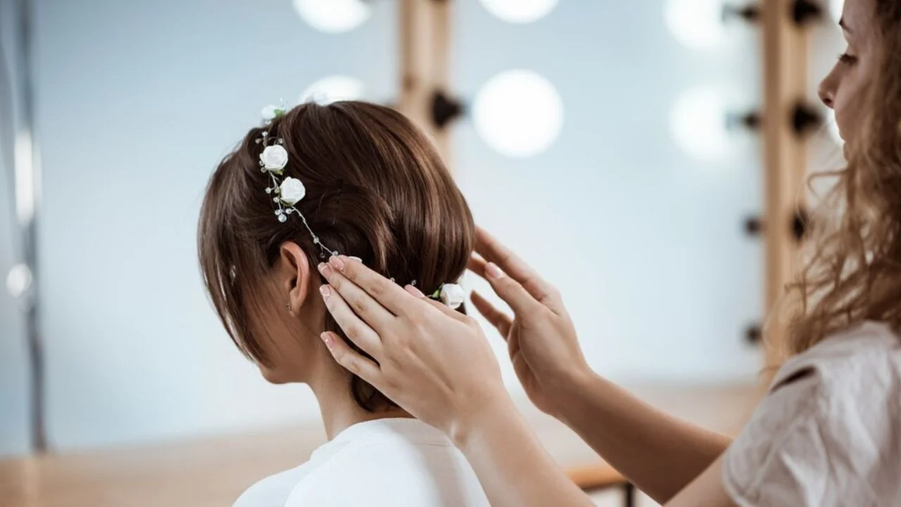 A hairstylist adjusting a delicate floral headband with small white flowers on a woman’s short brown hair, creating an elegant and vintage-inspired bridal look in a softly lit dressing room.