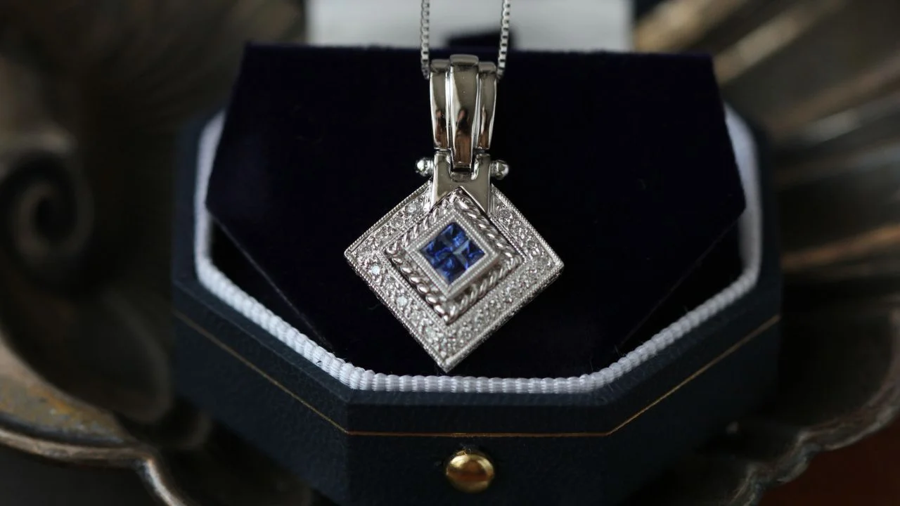 A stunning sapphire pendant set in a diamond-encrusted square frame, resting in a velvet-lined jewelry box.