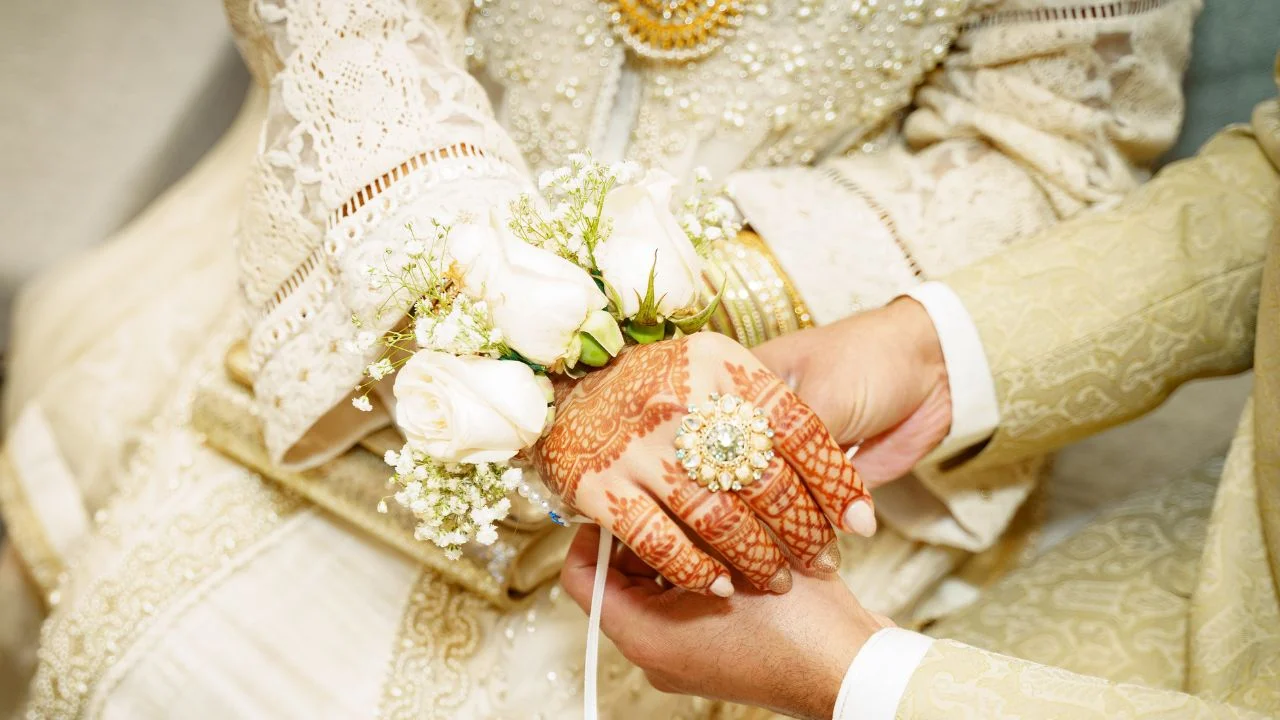 Bride wearing a white lace dress and has a beautiful mehndi designs on her hands. The groom is wearing a beige suit and is holding the bride's hand.