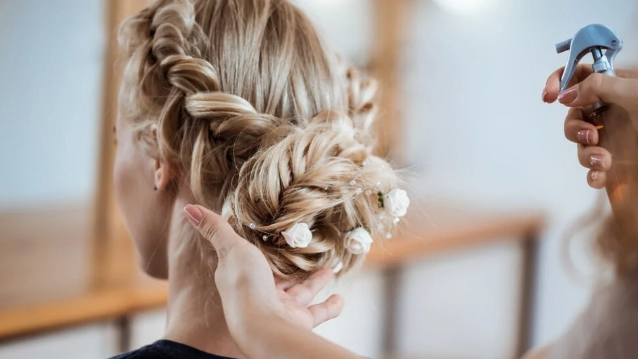 A hairstylist working on an intricate bridal updo with blonde hair, adorned with small white flowers, while applying hairspray to hold the style.