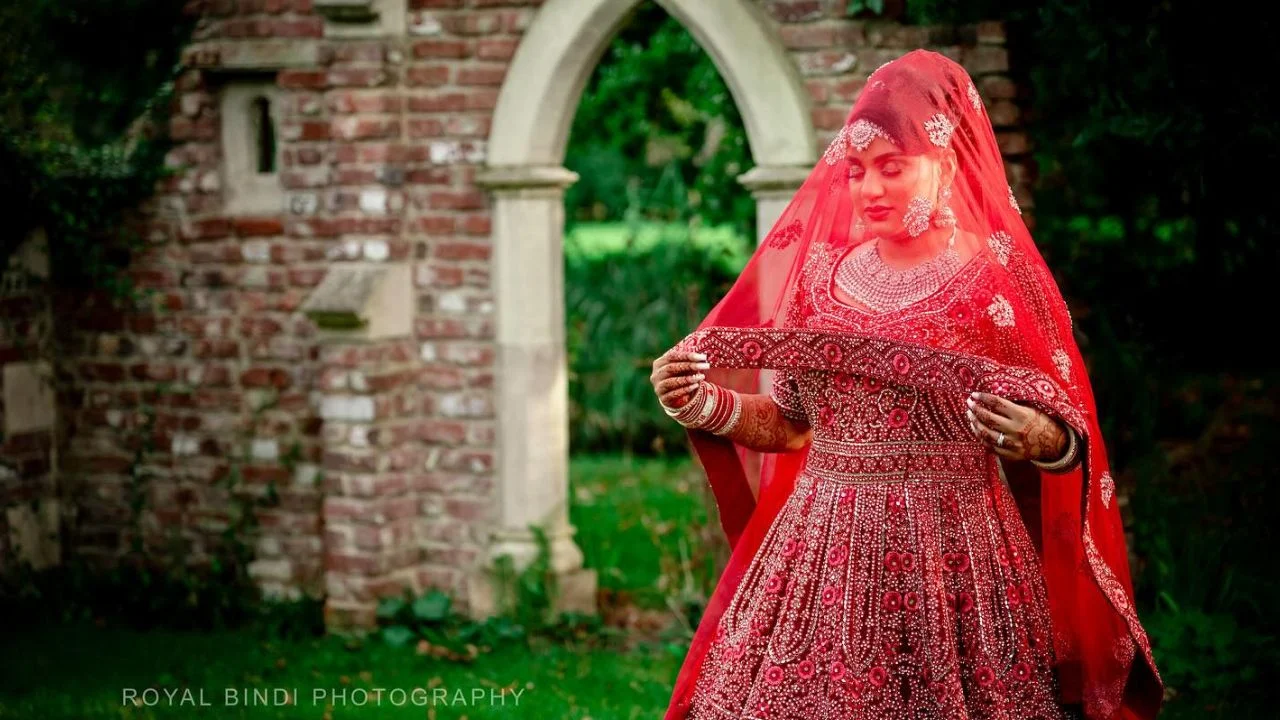 Indian bride in a red lengha stands in front of a brick wall wearing a red dupatta and holding the fabric of the veil in her hand.