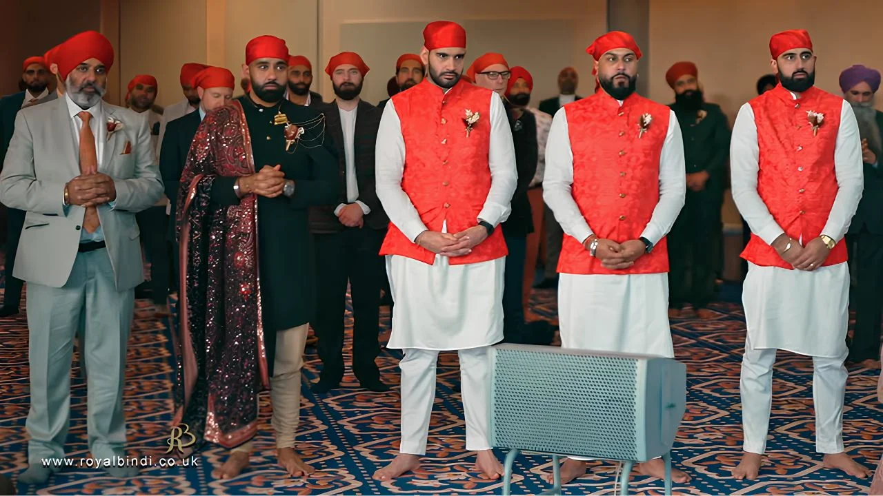 Groom and groomsmen dressed in traditional Sikh wedding attire with red turbans and orange vests, standing during a ceremony in a Sikh Gurdwara.