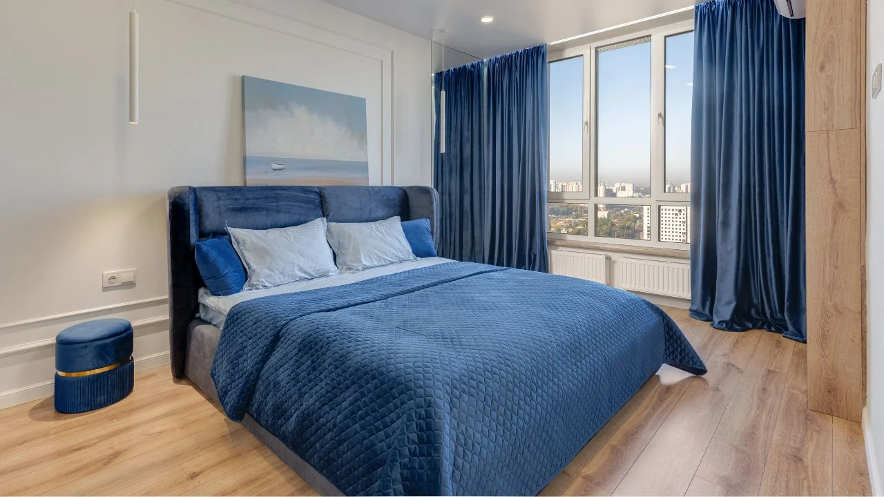 A modern bedroom featuring a blue bed, blue curtains, and a blue ottoman, with a wooden floor and a large window overlooking a city view.