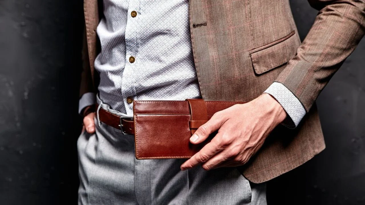 A man in a brown suit jacket and gray pants holds a brown leather wallet in his hand.