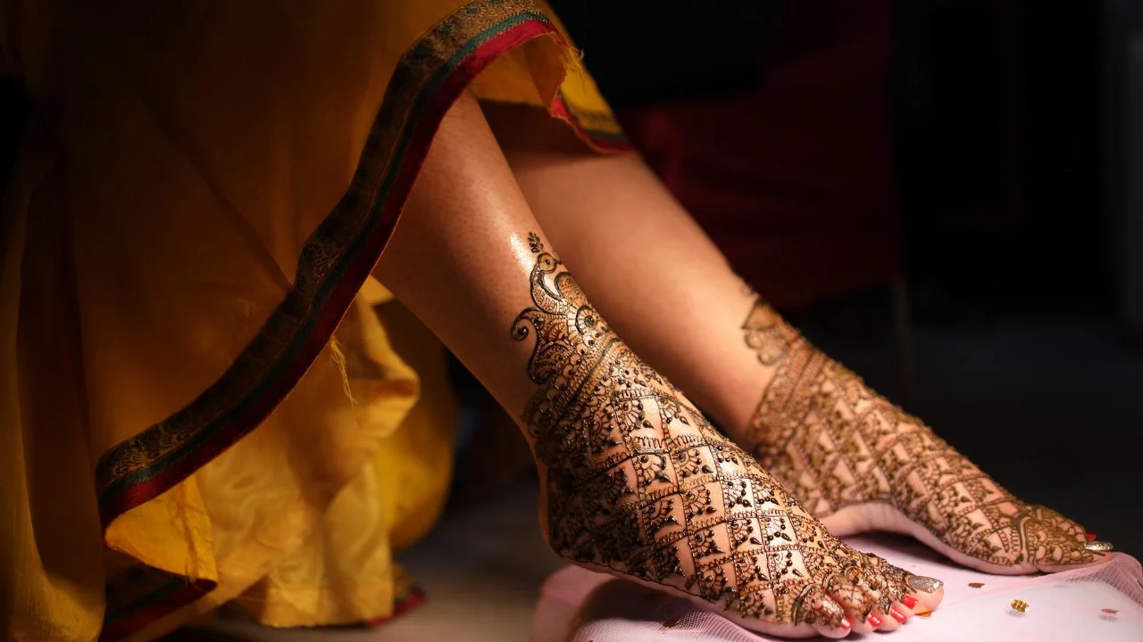 Woman's legs with intricate henna designs on her feet. The henna is dark brown and covers most of her ankles and feet. The woman is wearing a yellow sari with a red border.