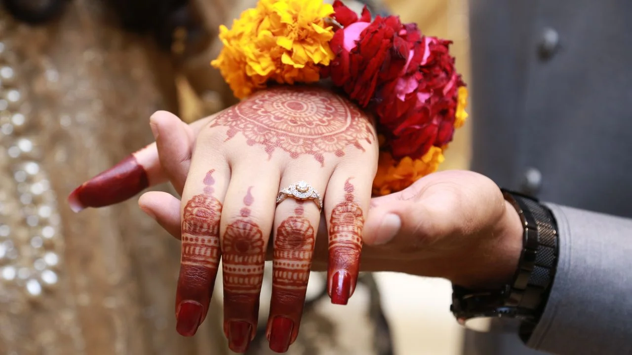 A close-up of a bride’s hand featuring an intricate mehndi design, adorned with a diamond ring and colourful floral decorations.