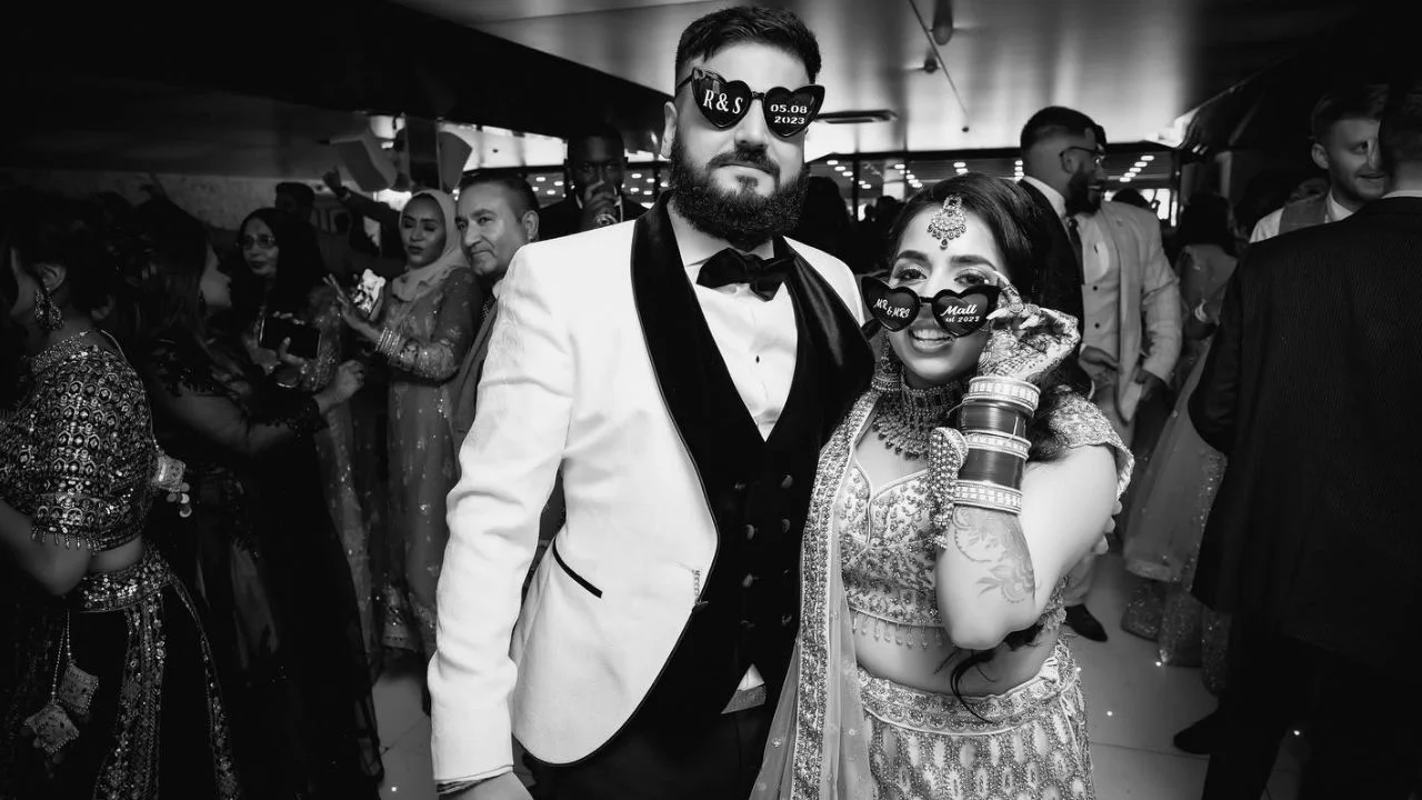 A black-and-white photo of a newlywed couple at their wedding reception. The groom is dressed in a white tuxedo jacket with a black bow tie, while the bride wears a traditional embellished outfit with intricate jewelry. Both are holding customized sunglasses with their wedding date and names on them, enjoying the moment amidst a festive crowd.