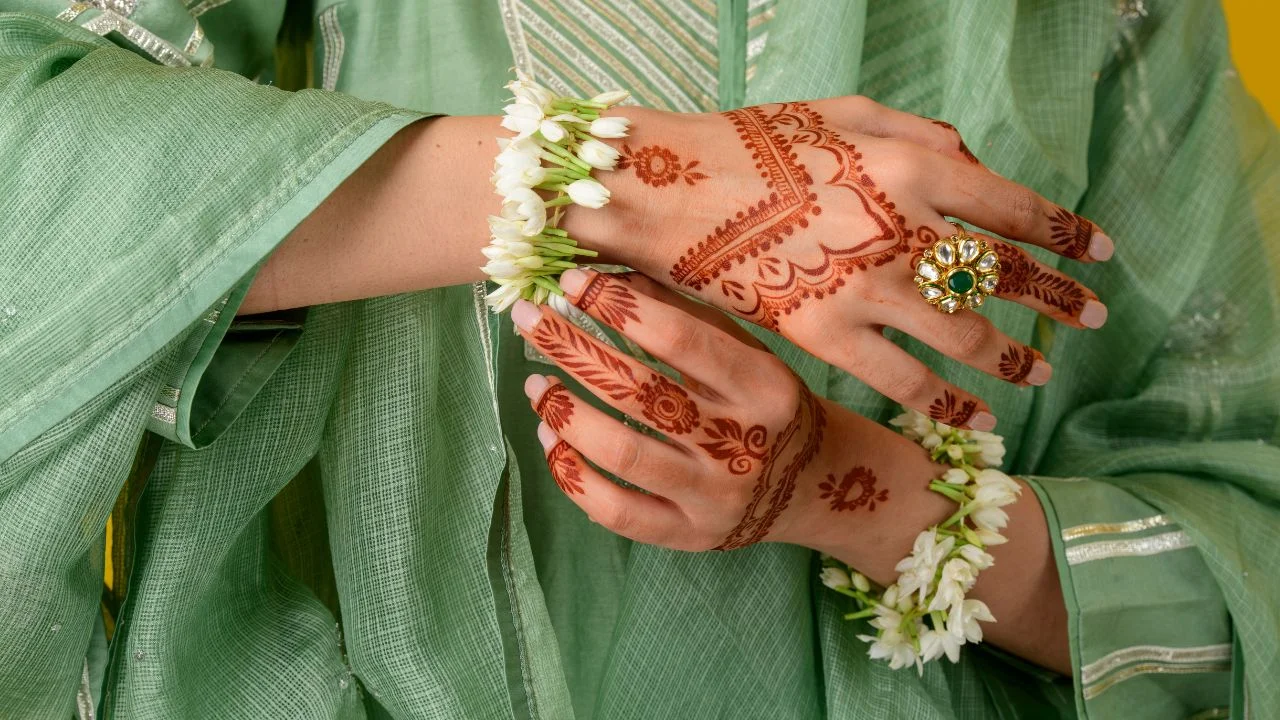 A close-up of hands featuring an open-space mehndi design, adorned with floral bracelets and a large statement ring, wearing a light green traditional outfit.