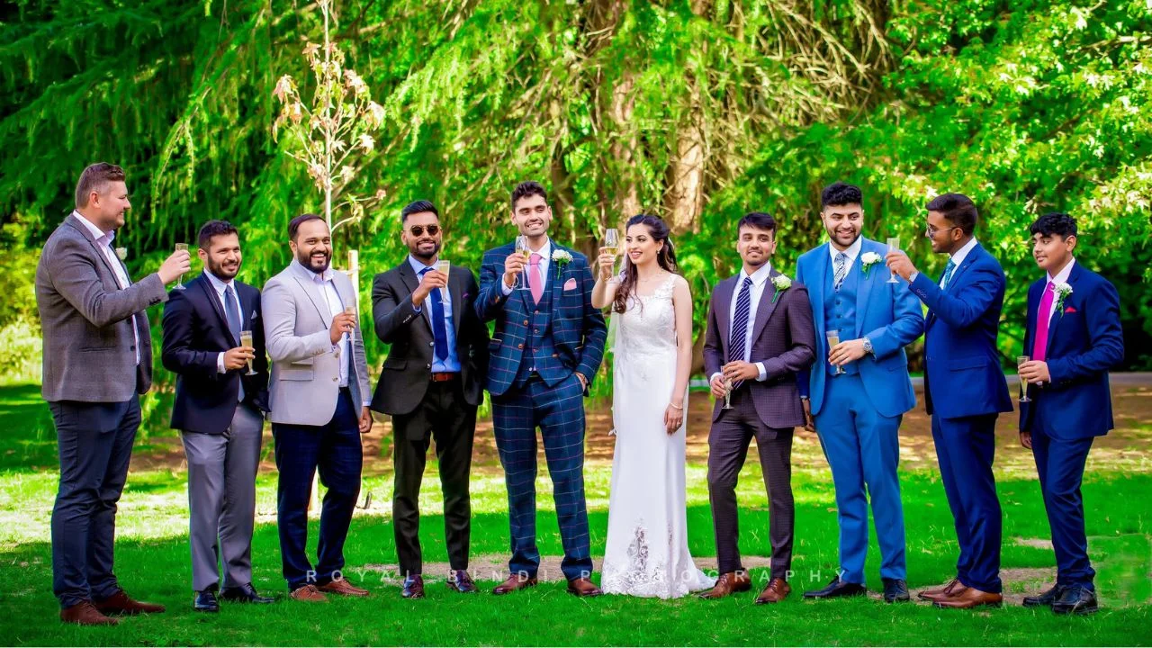 Bride, groom, and groomsmen enjoying a celebratory toast in an outdoor wedding setting with a lush green backdrop.