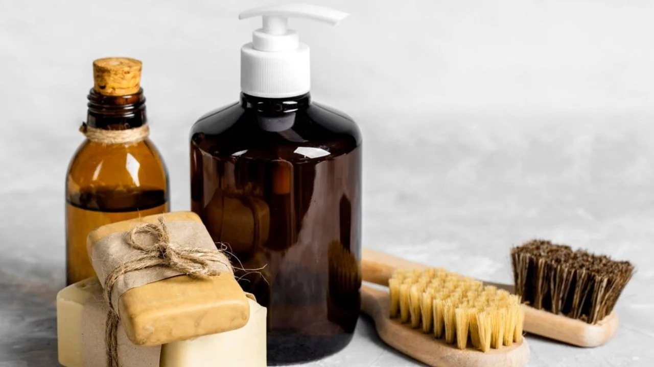 A collection of organic grooming products on a gray background. The products include a glass bottle of oil, a pump dispenser bottle of liquid soap, two bars of wrapped soap, and a wooden brush.