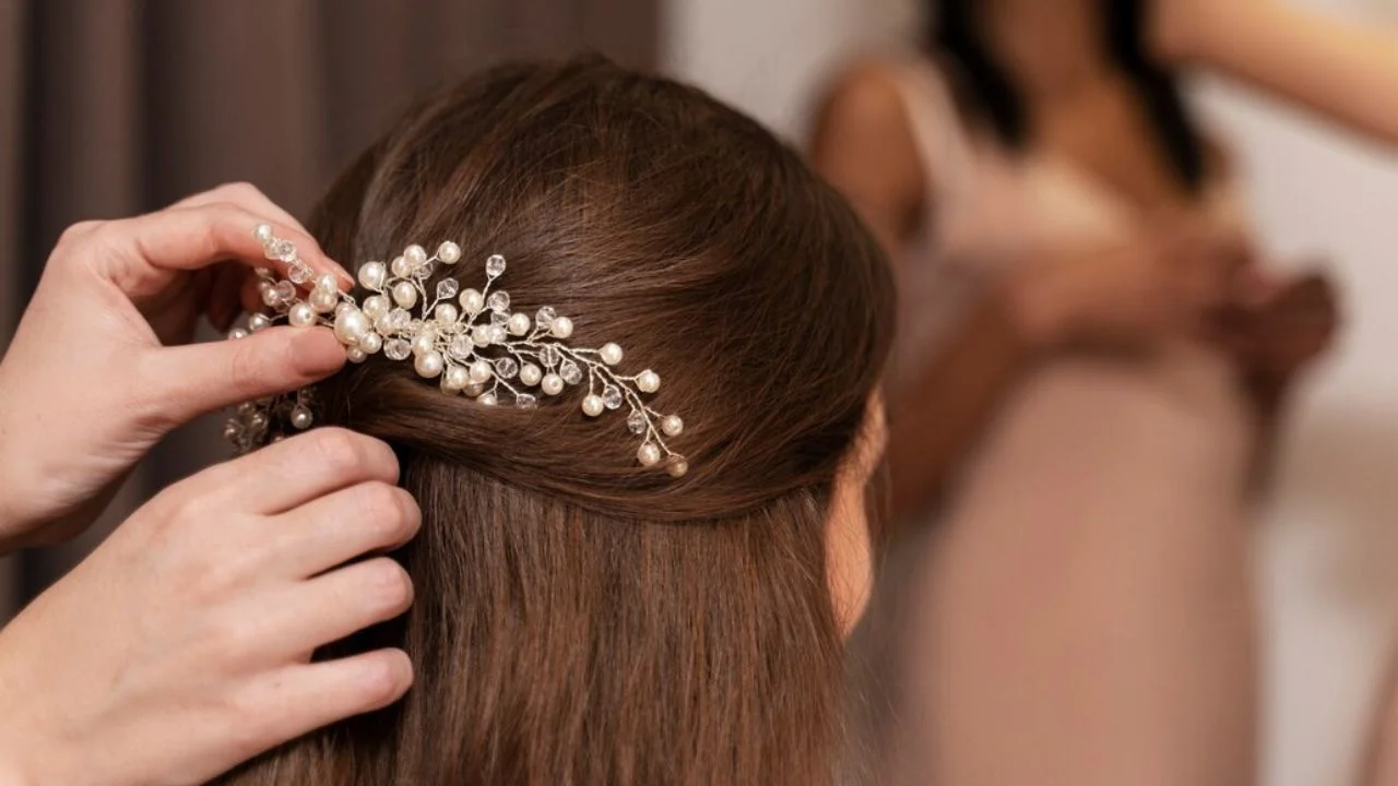 A close-up of hands placing a pearl-studded decorative hair clip into a bride's brown hair, with a blurred figure in the background.