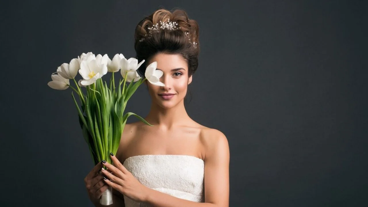 A bride in a strapless white dress holding a bouquet of white tulips in front of her face, with a pearl-embellished hairpiece in an elegant updo against a dark background.