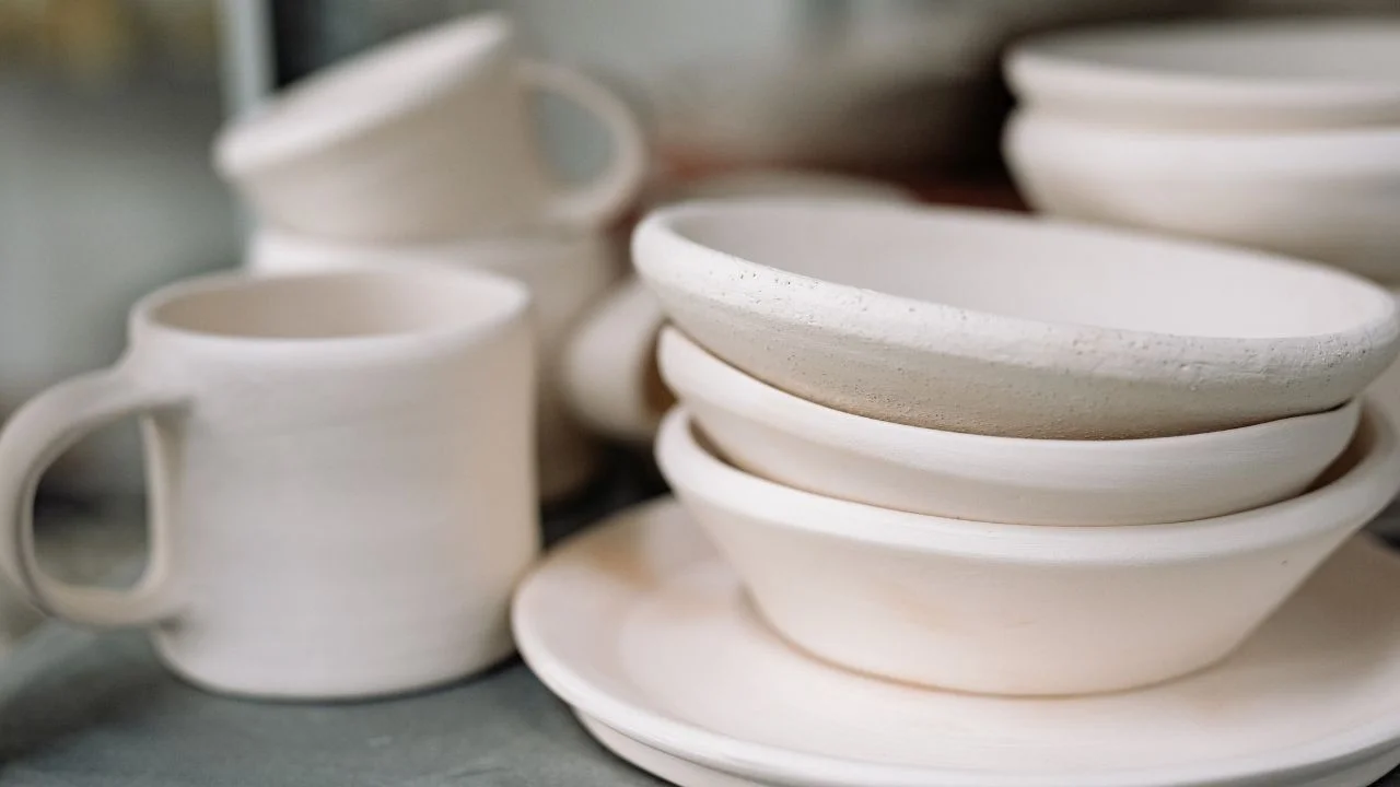 A collection of handmade ceramic pottery, including mugs, bowls, and plates, stacked and displayed on a shelf.