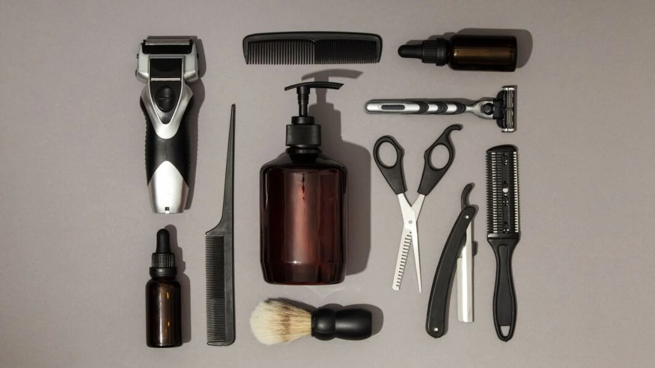 A flat lay of various men's grooming tools on a gray background, including an electric razor, a shaving brush, a comb, scissors, a straight razor, and a bottle of beard oil.