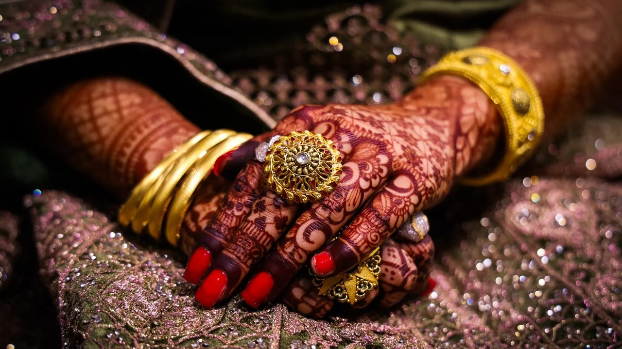 Close up of a woman's hands with intricate henna designs and gold jewelry, holding her hands together. The background is a blurred out fabric with sequins.