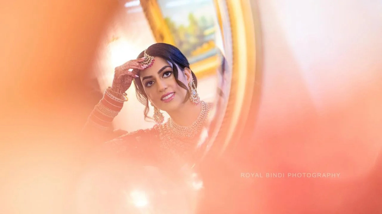 A bride in traditional Indian attire adjusts her jewellery in front of a mirror.