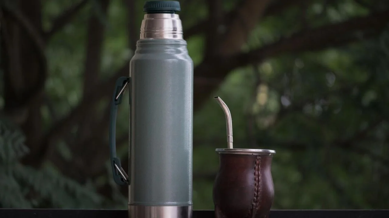 A gray thermos and a brown leather mate gourd with a metal bombilla straw are sitting on a table with a blurred green background.