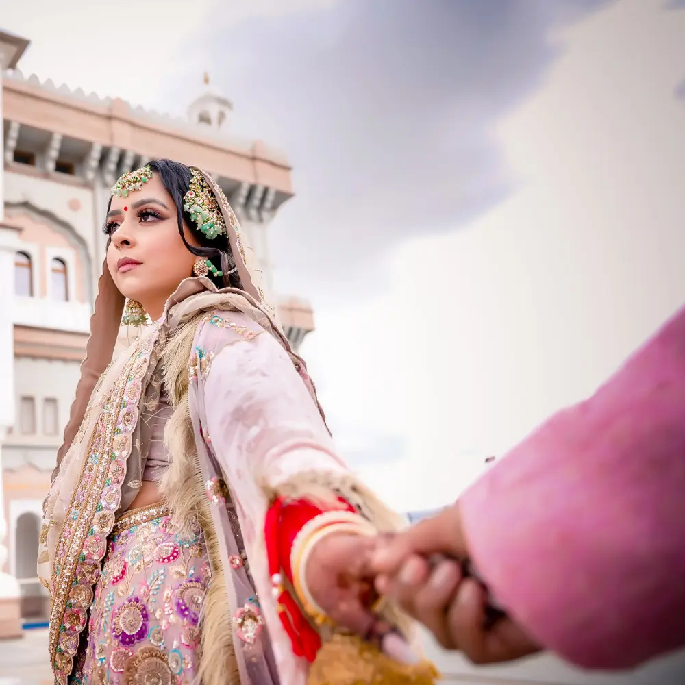 A Sikh bride in intricately detailed traditional wedding attire, holding hands with her partner outside a stunning architectural backdrop, captured by Royal Bindi photography.