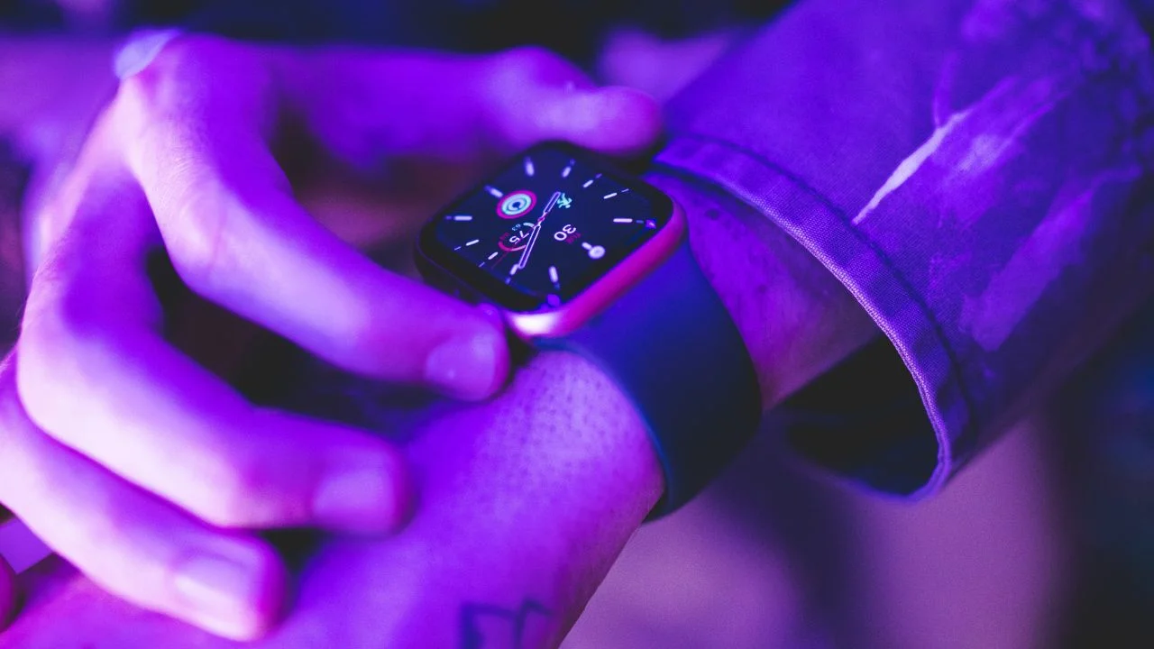 A close-up photo of a person's hand swiping on a Smart Watch with a purple strap. The watch face shows the time and various app icons