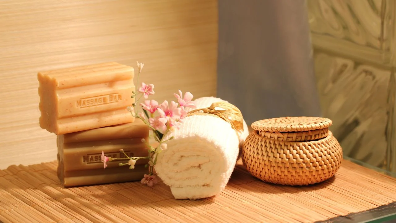 A spa treatment setup featuring handmade soaps, a rolled-up white towel, and a small woven basket with flowers.