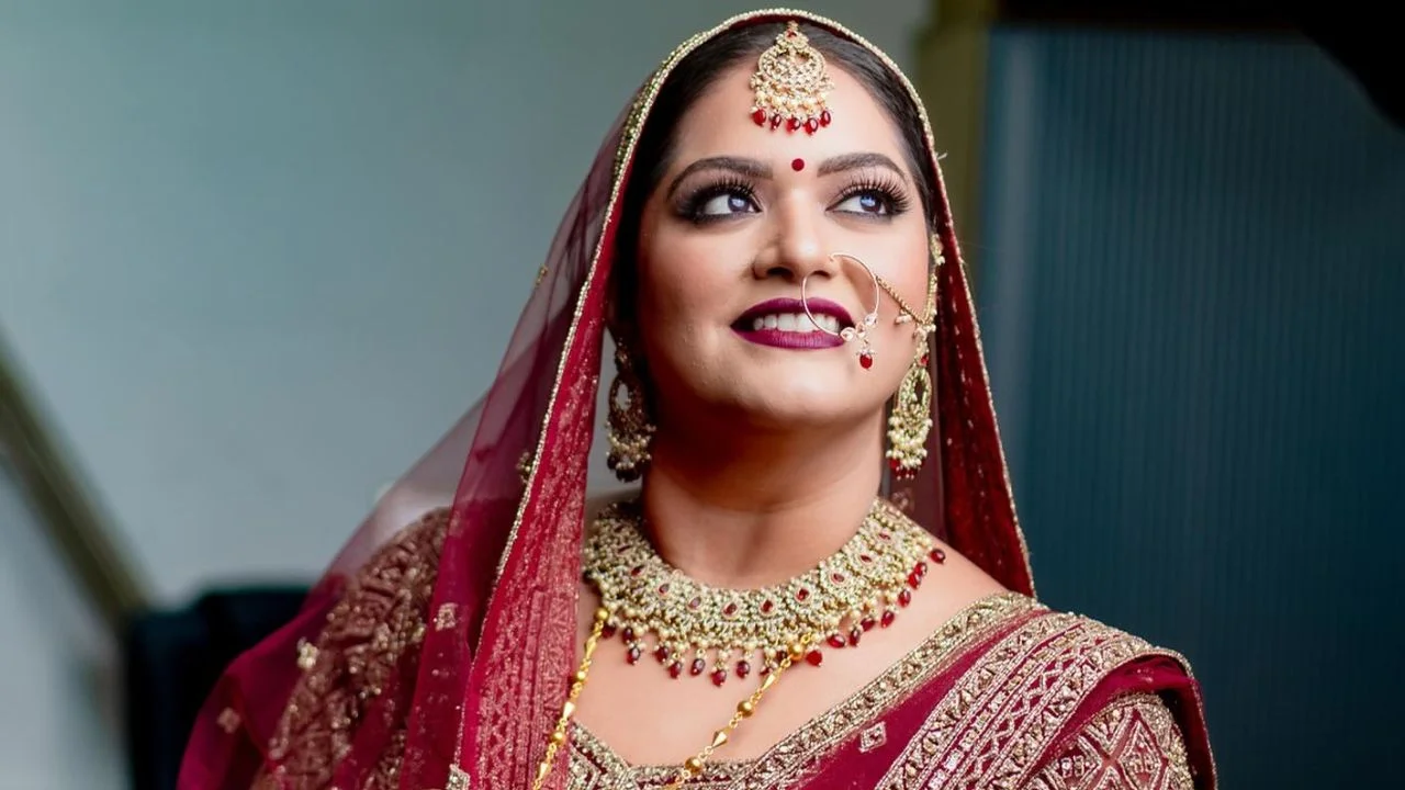 A young woman in a red saree with a gold necklace and other jewellery, including a nose ring. She is looking off to the side with a slight smile on her face.