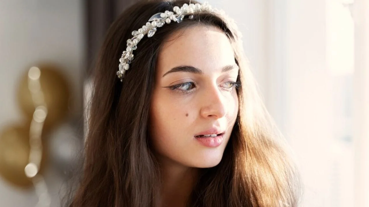 A bride with long brown hair wearing a pearl and crystal-embellished headband, looking to the side in soft natural light, with a blurred background.