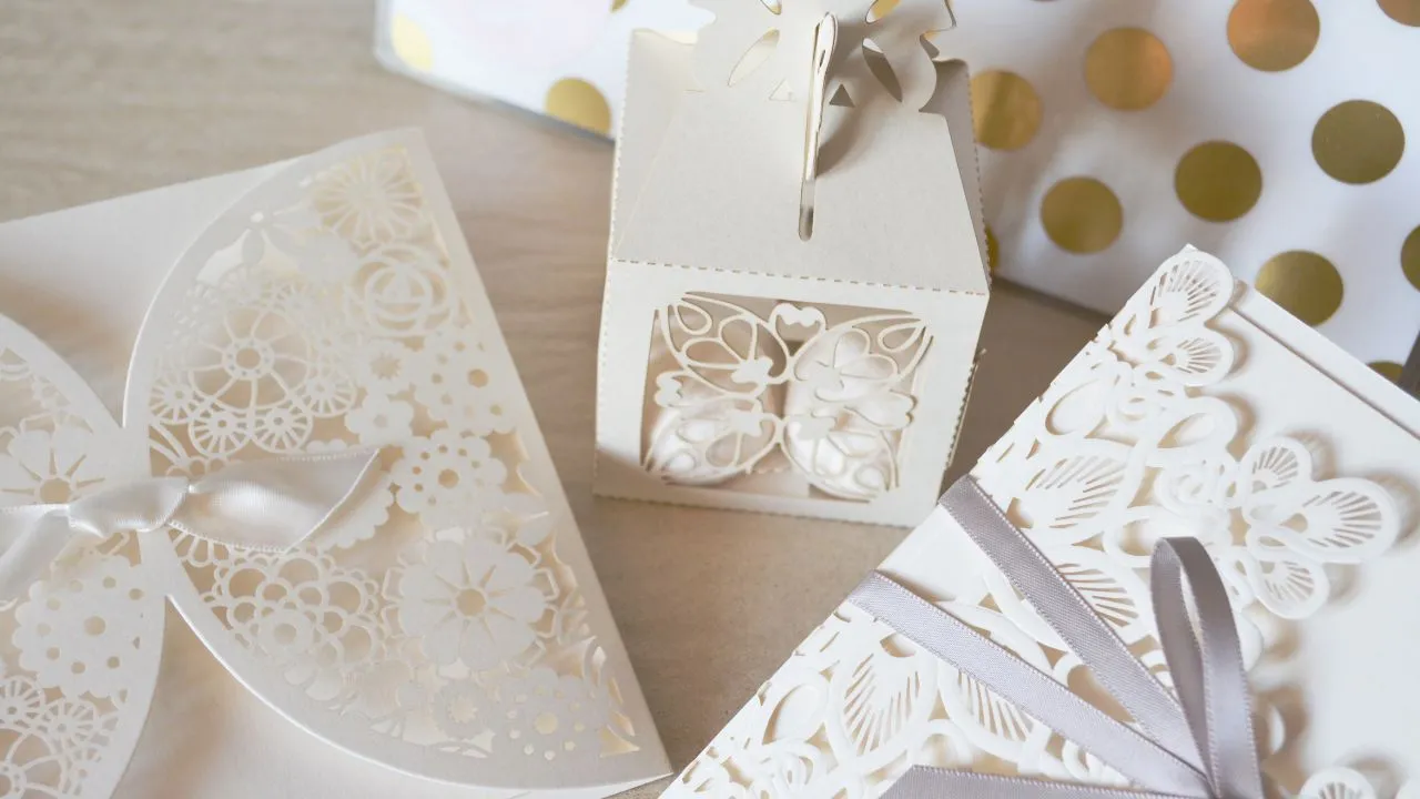 A close-up photo of three white wedding invitations with intricate laser-cut patterns and ribbons. One invitation is folded with a bow, another is in a box with a lid, and the third is open with a ribbon tied around it.