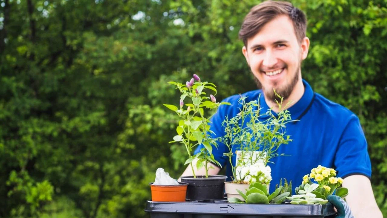 A smiling person in a blue shirt holding a tray filled with small potted plants, set against a backdrop of lush greenery.