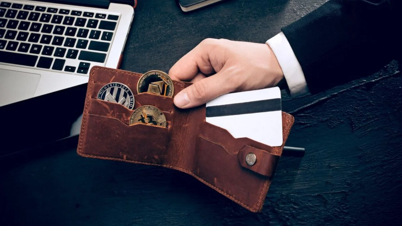 A man in a suit is holding open a brown leather wallet. Inside the wallet are multiple cryptocurrency coins and a credit card. A laptop is visible on the desk in the background.