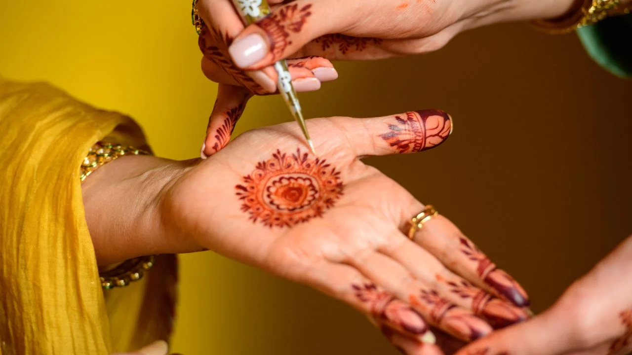 A close-up of a person's hand applying henna to another person's hand. The henna paste is dark brown and is being applied with a small tool.