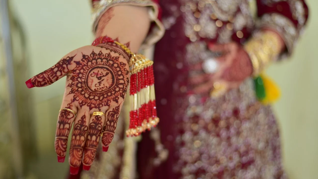 A close-up of a woman's hand adorned with intricate henna designs. The henna is dark brown and covers most of her palm and fingers.