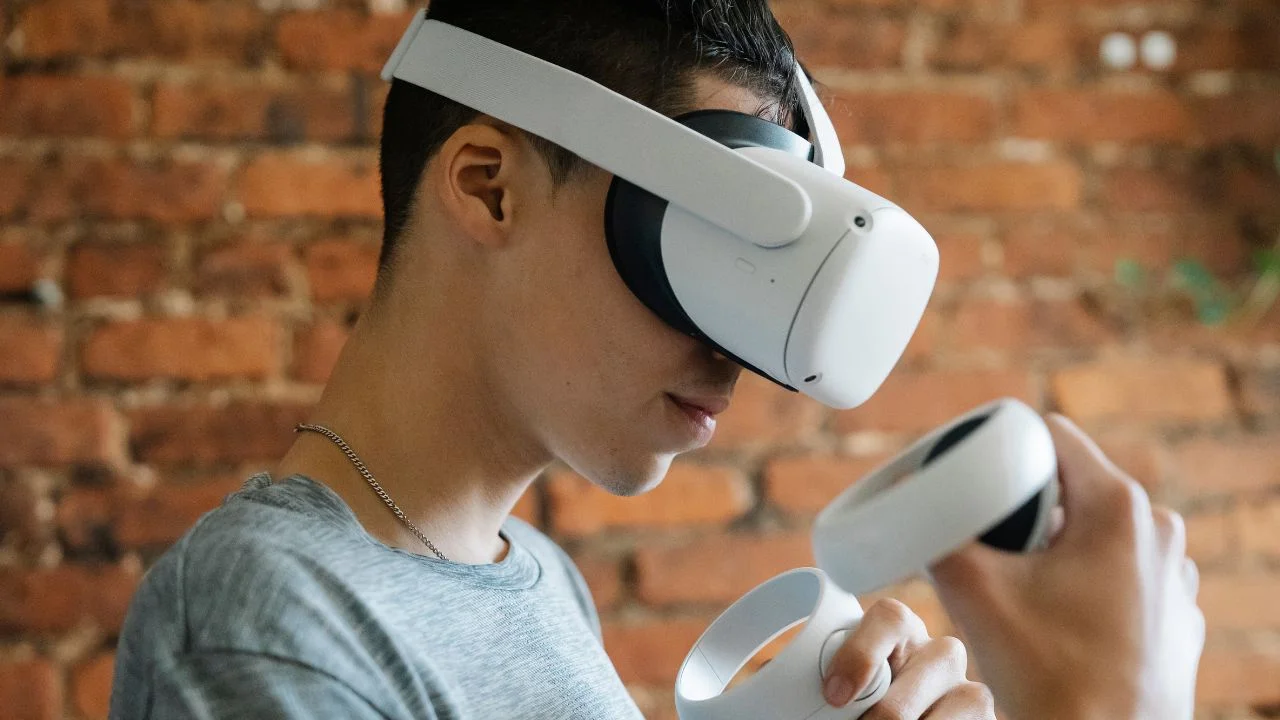 Person wearing a virtual reality headset and holding controllers, engaging in a VR experience against a brick wall background.