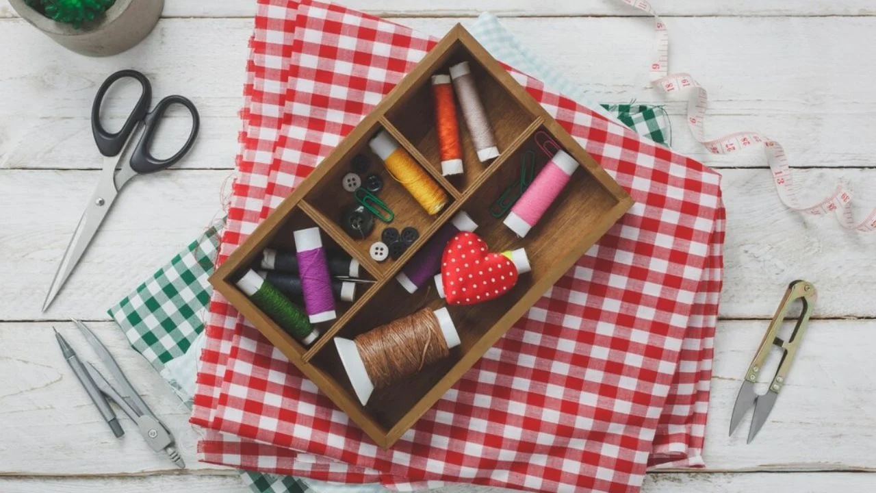 A wooden box filled with various sewing supplies, including thread, buttons, scissors, and a measuring tape, surrounded by different fabrics on a white wooden table.