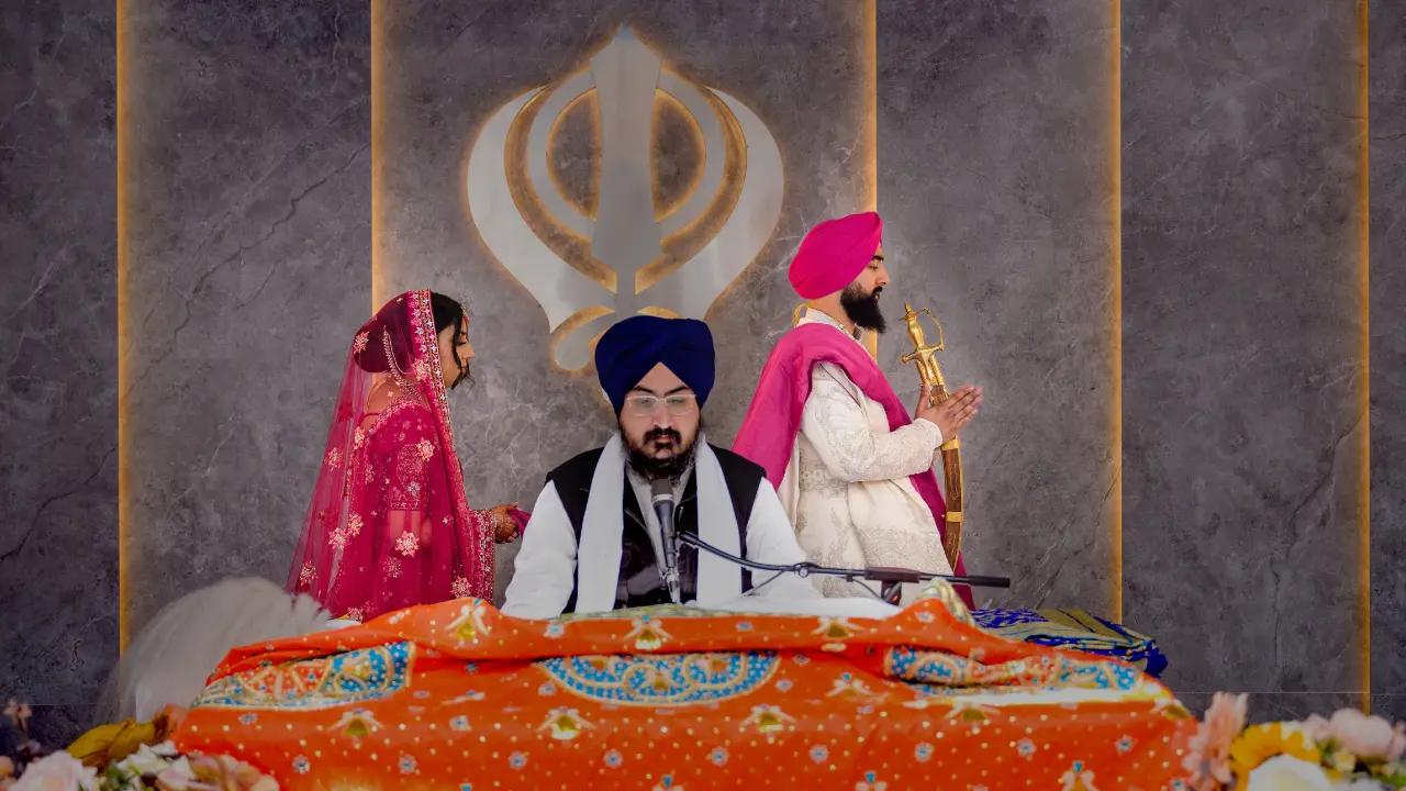 A Sikh wedding ceremony is taking place. The bride and groom are standing in front of the altar, while a priest is reading from a book. The ceremony is being held in a temple, and the walls are decorated with traditional Sikh symbols.