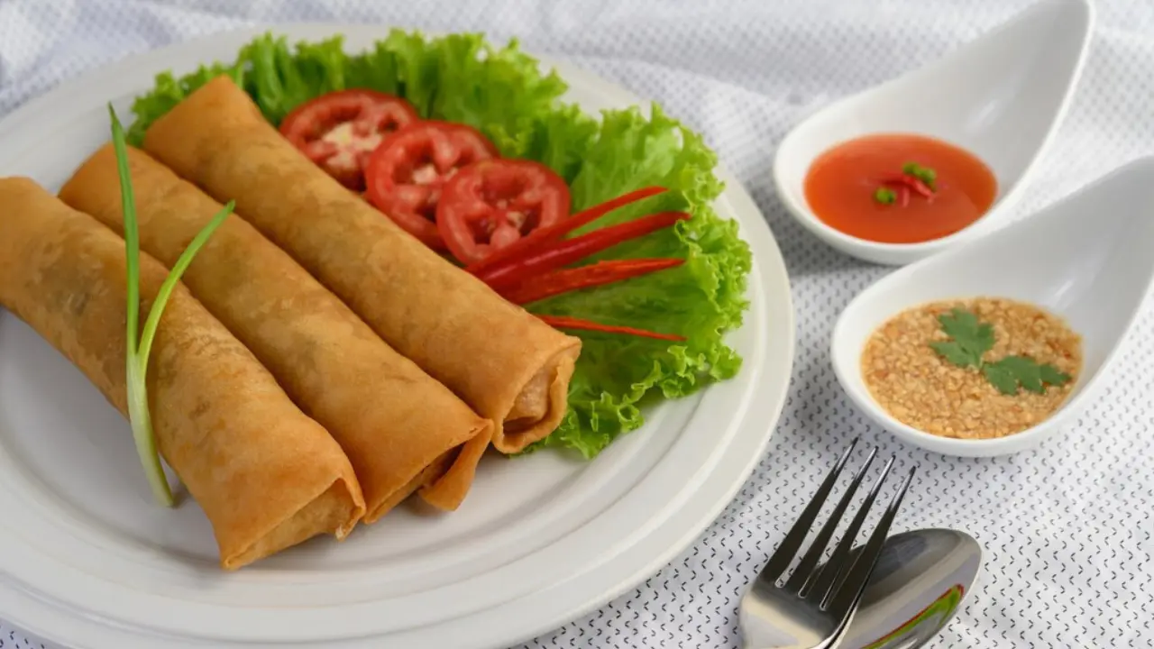 Three golden brown spring rolls on a white plate with lettuce, tomatoes, chili peppers, and dipping sauces.